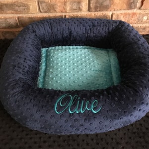 May include: A navy blue pet bed with a teal interior. The bed has a plush, textured exterior and features the name "Olive" embroidered in teal thread. The bed is designed with a raised rim for added comfort and support.