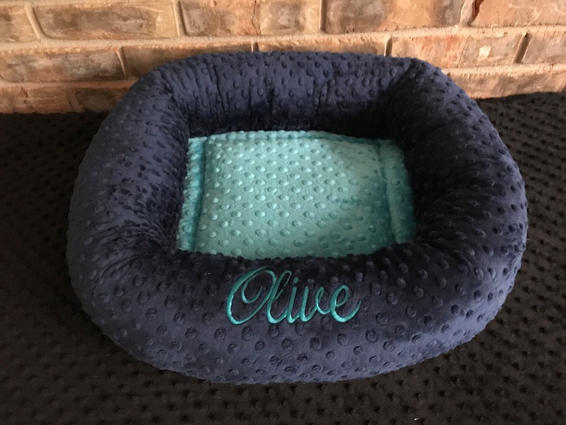 May include: A navy blue pet bed with a teal interior. The bed has a plush, textured exterior and features the name "Olive" embroidered in teal thread. The bed is designed with a raised rim for added comfort and support.