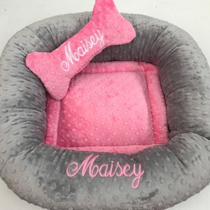 May include: A gray and pink dog bed with a bone-shaped pillow. The bed has a gray, textured exterior and a pink interior. The name "Maisey" is embroidered on both the bed and the pillow in pink thread.