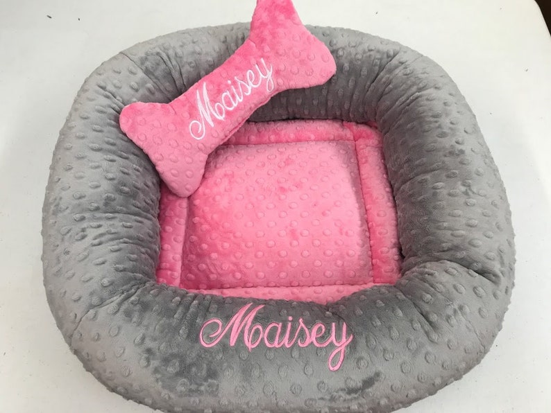 May include: A gray and pink dog bed with a bone-shaped pillow. The bed has a gray, textured exterior and a pink interior. The name "Maisey" is embroidered on both the bed and the pillow in pink thread.