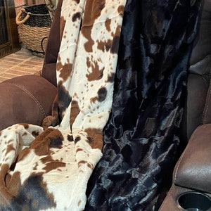 May include: Two plush blankets, one with a brown and white cow print and the other black, are draped over a brown leather recliner.
