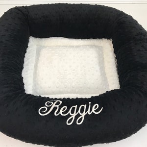 May include: Black and white pet bed with the name "Reggie" embroidered in white script. The bed has a plush, textured black exterior and a soft, white interior with a dotted pattern. The bed is designed for comfort and support.