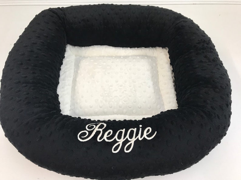 May include: Black and white pet bed with the name "Reggie" embroidered in white script. The bed has a plush, textured black exterior and a soft, white interior with a dotted pattern. The bed is designed for comfort and support.