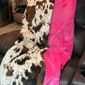 May include: A cozy blanket with a cowhide pattern in brown, white, and black, paired with a vibrant pink blanket. The blankets are draped over a brown leather recliner, creating a warm and inviting aesthetic.