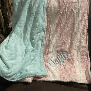 May include: A soft, plush blanket with a light blue side and a pink side. The pink side has the name "Taylor" embroidered on it in a light blue thread.