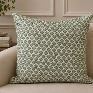 May include: A square throw pillow featuring a repeating pattern of small, sage green, heart-shaped motifs on a cream-colored fabric background. The pillow has a structured, plump appearance with clean, finished edges and a consistent, dense textile weave.