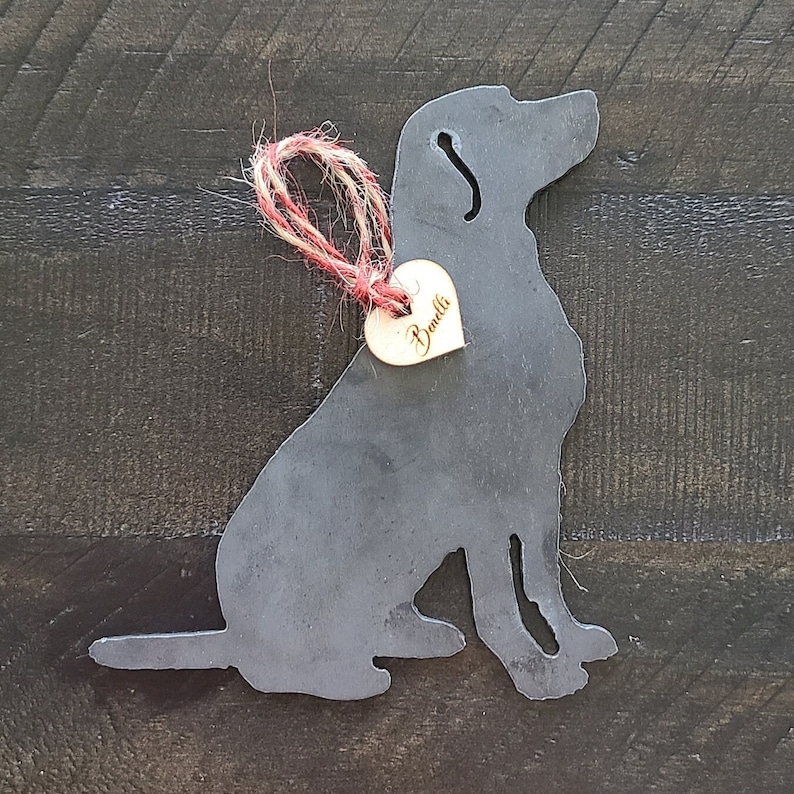 May include: A black metal silhouette of a dog sitting, with a small wooden heart-shaped tag that says "Bandit" hanging from a red and white twine.