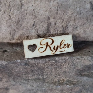 May include: A wooden sign with the name "Ryler" engraved in cursive script. A small heart is carved into the wood to the left of the name.