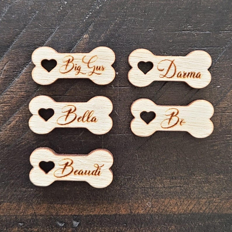 May include: Five wooden dog bone-shaped tags with heart cutouts and personalized names: Big Gus, Darma, Bella, Be, and Beaudi.