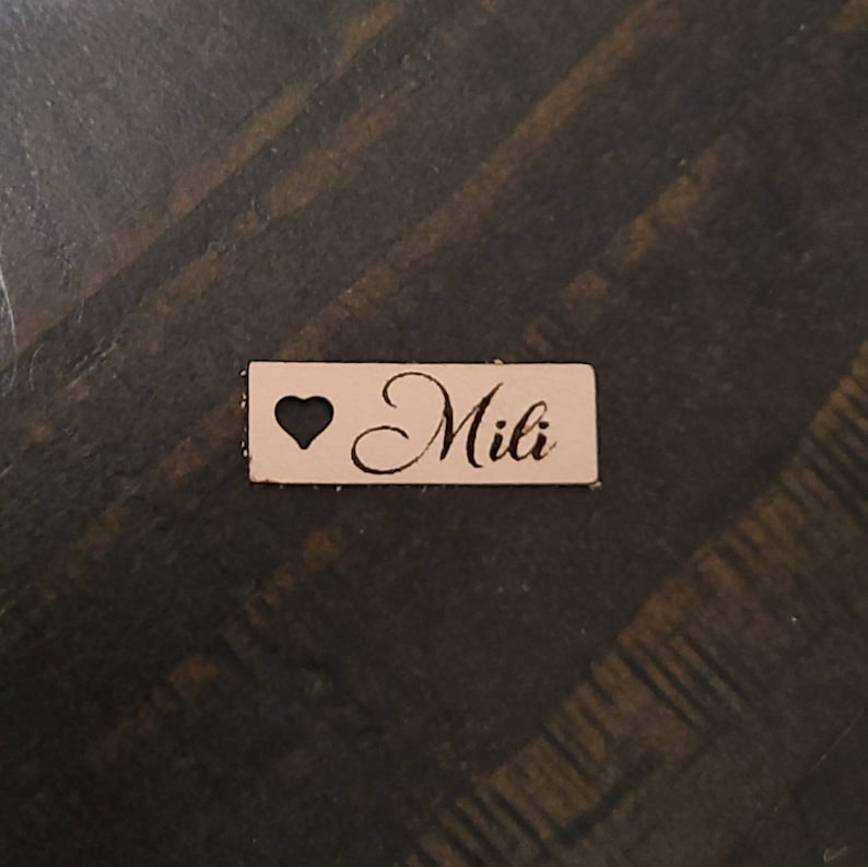 May include: A light pink wooden tag with a heart cutout and the name "Mili" engraved in cursive.