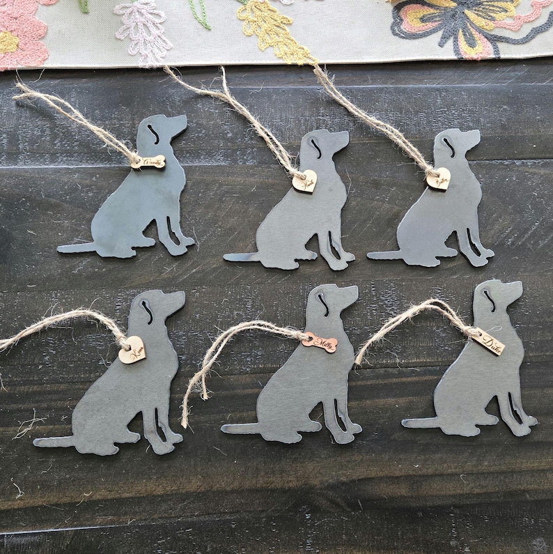 May include: Six metal ornaments shaped like labrador retrievers with a small metal tag attached to each. The tags have different names on them, including "Max", "Duke", and "Mia".
