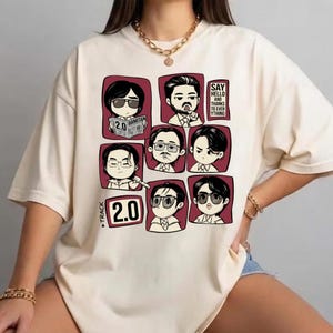 May include: Cream-colored t-shirt featuring a grid of cartoon illustrations of people with sunglasses and the text "2.0" and "Say Hello and Thanks to Ever Ything". The shirt has a crew neck and short sleeves.