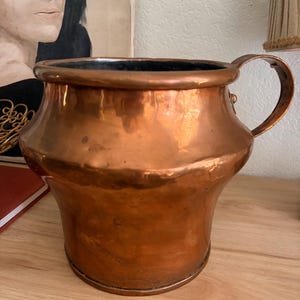 May include: A polished copper pot with a wide, rounded body, a thick rim, and a single curved handle attached to the side. The exterior surface shows a warm, metallic orange-brown finish with subtle variations in texture and light reflection.