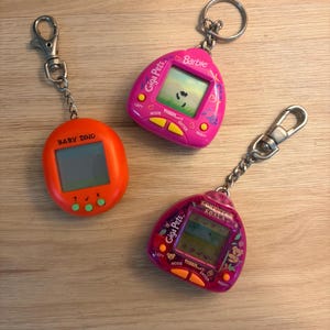 May include: Three digital pet keychains: an orange Baby Dino device, a pink Giga Pets Barbie device, and a dark pink Giga Pets Komputer Koala device. Each unit features a small LCD screen, control buttons, and a metal chain with a lobster clasp.