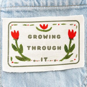 May include: A light blue denim pocket featuring a rectangular white fabric patch. The patch displays the text "GROWING THROUGH IT" in dark green letters, surrounded by an embroidered border with red tulips and green stems and leaves.