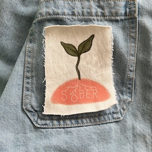 May include: A light blue denim pocket with a white patch. The patch shows a plant growing from a pink base, with the word "SOBER" in pink. The plant has green leaves and a brown stem.