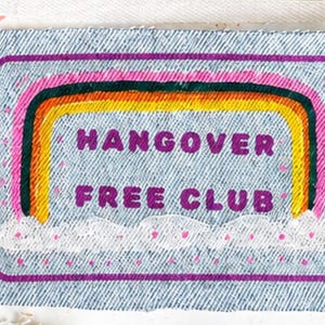 May include: A rectangular denim patch featuring a painted rainbow with pink, orange, yellow, and dark green bands. Below the rainbow are white clouds with pink dots. The text HANGOVER FREE CLUB is printed in purple letters in the centre.