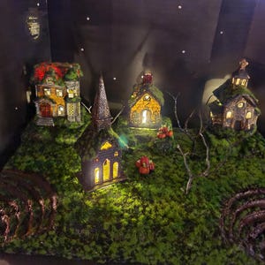 May include: A miniature village scene with four houses, each with a lit window, set on a bed of green moss. The houses are made of wood and have a rustic, whimsical style. The scene is lit by small lights, creating a warm and inviting atmosphere.