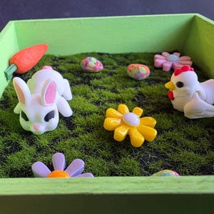 Op de afbeelding: Een groene houten kist gevuld met kunstgras, met paasfiguren. Inclusief een witte konijn met roze oren, een wortel, een witte kip, kleurrijke eieren en gele en paarse bloemen.