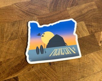 May include: A sticker in the shape of Oregon with a sunset scene. The design features a yellow sun, a dark mountain silhouette, two surfboards, and the words "Pacific City OR" in a stylized font. The sky transitions from blue to orange.