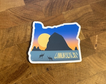 May include: A sticker in the shape of Oregon with a sunset scene featuring a large rock formation, a yellow sun, and two deer. The text "Cannon Beach" is printed in white.