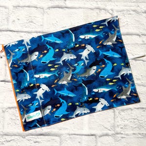 May include: A rectangular fabric pouch featuring a dark blue background with a repeating pattern of various shark species and small yellow fish. The pouch has an orange zip along the side and a small white label with the text Bebeco printed on it.