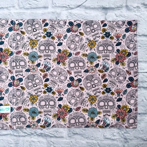 May include: A rectangular fabric pouch featuring a repeating pattern of black-outlined skulls and colourful flowers on a light pink background. The pouch includes a pink zip along the left side and a small white label with the text Boboco.