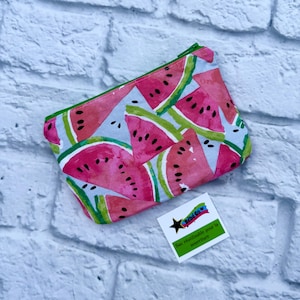 Könnte beinhalten: Eine rechteckige Reißverschlusstasche mit einem lebendigen Wassermelonenmuster in Rosa-, Grün- und Weißtönen auf hellblauem Grund. Ein kleines weißes quadratisches Etikett ist beigefügt, das ein schwarzes Sternlogo und den Text Bébé Co. zeigt.
