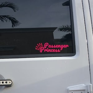 May include: A white Jeep with a black and white American flag decal on the back window. The back window also has a pink decal that says "Passenger Princess" with a crown above the word "Passenger".