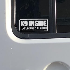 May include: A rectangular white sticker with black text that reads "K9 INSIDE TEMPERATURE CONTROLLED" is affixed to a vehicle window. The sticker is in a black rectangular border. The vehicle is white.