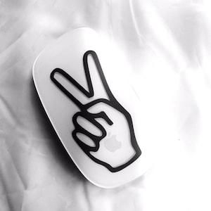 May include: A white wireless mouse with a black peace sign sticker on the top. The peace sign is a hand with two fingers raised in the air.