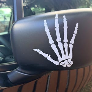 May include: Black side mirror with a white skeleton hand decal. The hand is in a flat, stylized design, with all the bones visible. The mirror is attached to a black vehicle.