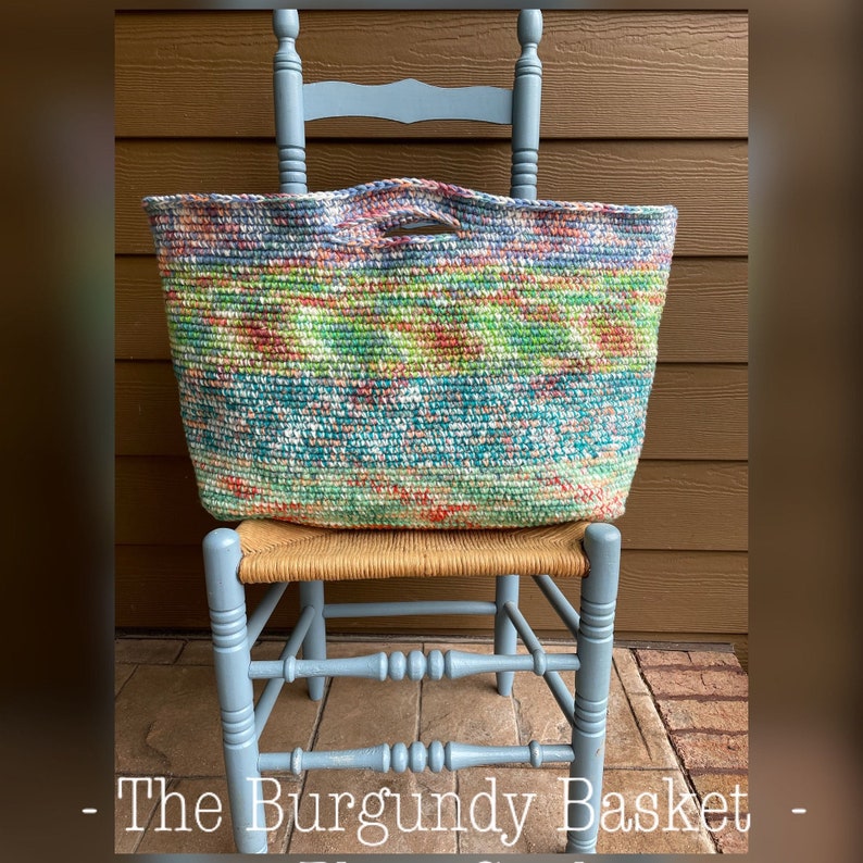 Pu&ograve; includere: Una borsa a mano in crochet colorata con un manico intrecciato &egrave; appoggiata su una sedia di legno blu chiaro. La borsa ha un motivo multicolore con sfumature di verde, blu, rosa e arancio. Il testo "The Burgundy Basket" &egrave; visibile nella parte inferiore dell'immagine.