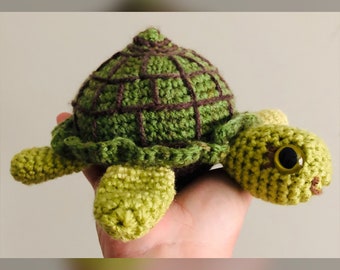 May include: A crocheted green turtle with brown accents and large black eyes. The turtle is sitting on a hand.
