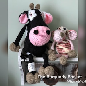 Könnte beinhalten: Eine schwarz-weiße gehäkelte Kuh mit rosa Akzenten sitzt auf einem Regal neben einem kleineren gehäkelten Schwein mit rosa und braunen Akzenten. Die Kuh hat ein Schild mit der Aufschrift "Crocheted with love".