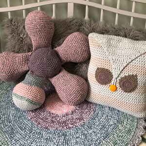 May include: Two crocheted pillows, one shaped like a flower in shades of pink and gray, and the other shaped like an owl in shades of beige, gray, and brown, are sitting on a gray and white crocheted blanket.
