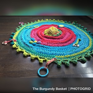 Könnte beinhalten: Eine bunte gehäkelte Spielmatte mit einem kreisförmigen Design in Grün-, Blau-, Rosa- und Gelbtönen. Die Matte hat verschiedene Texturen und ist mit Holz- und Plastikspielzeug ausgestattet.