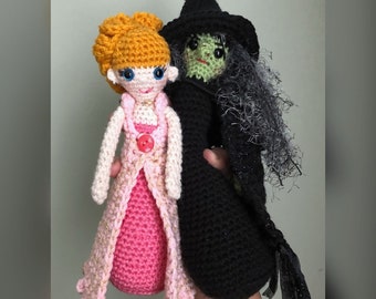 May include: A crocheted doll with long blonde hair wearing a pink dress and a crocheted witch doll wearing a black dress and a pointed hat. The witch doll has green skin and black hair.