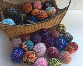 May include: A collection of round, crocheted spheres in various colors and textures, including solid, variegated, and fuzzy yarns. Some spheres are contained within a woven wicker basket, while others are arranged on a flat surface in front.
