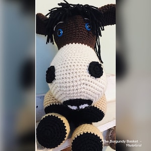 May include: A crocheted stuffed animal donkey with brown and tan fur, black hooves, and blue eyes. The donkey is sitting on a white shelf.