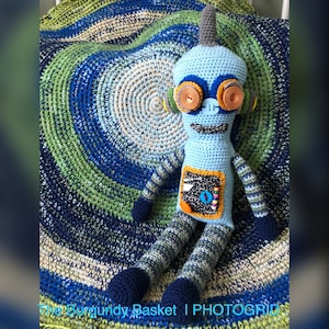 May include: A handmade, light blue crochet robot doll with orange and brown accents. The robot has striped arms and legs, a gray antenna, and a black and white panel on its chest. It is sitting on a blue, green, and white crocheted blanket.