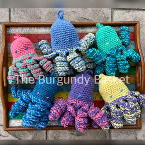 May include: Six crocheted octopus plushies in various colors, including pink, blue, green, purple, and yellow. The plushies are arranged on a wooden tray with a ruler.