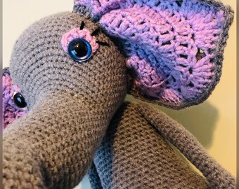 Op de afbeelding: Een grijze gehaakte olifant met paarse gehaakte oren. De olifant heeft grote blauwe ogen en een roze neus.
