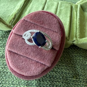 May include: A silver ring with a large, oval, dark blue gemstone, surrounded by smaller clear stones. The ring rests on a pink velvet ring box, with a green velvet jewelry box in the background.