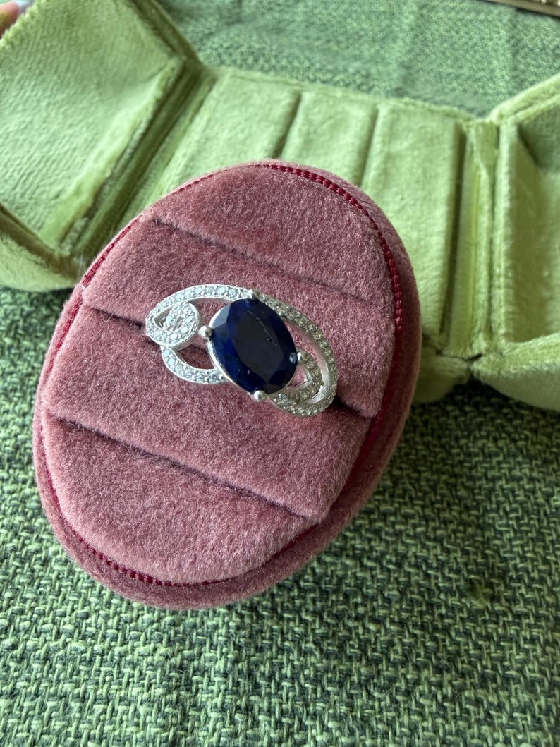 May include: A silver ring with a large, oval, dark blue gemstone, surrounded by smaller clear stones. The ring rests on a pink velvet ring box, with a green velvet jewelry box in the background.