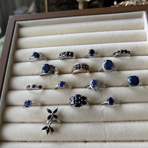 May include: A display of various sapphire rings in a wooden jewelry tray. The rings feature different designs, including solitaire, cluster, and band styles, with silver and gold settings. The sapphires are a deep blue color.