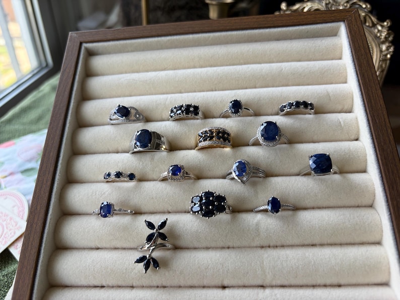 May include: A display of various sapphire rings in a wooden jewelry tray. The rings feature different designs, including solitaire, cluster, and band styles, with silver and gold settings. The sapphires are a deep blue color.
