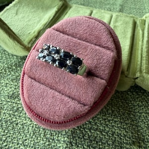 May include: A silver ring with two rows of oval, dark blue gemstones and small, clear stones. The ring is displayed in a pink velvet ring box with a red zipper, set against a green textured background.