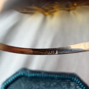 May include: A gold-toned bracelet with the engraved letters "ADLI" is displayed. The bracelet is slightly curved and rests above a teal, hexagonal jewelry box. The background is a soft, blurred white.