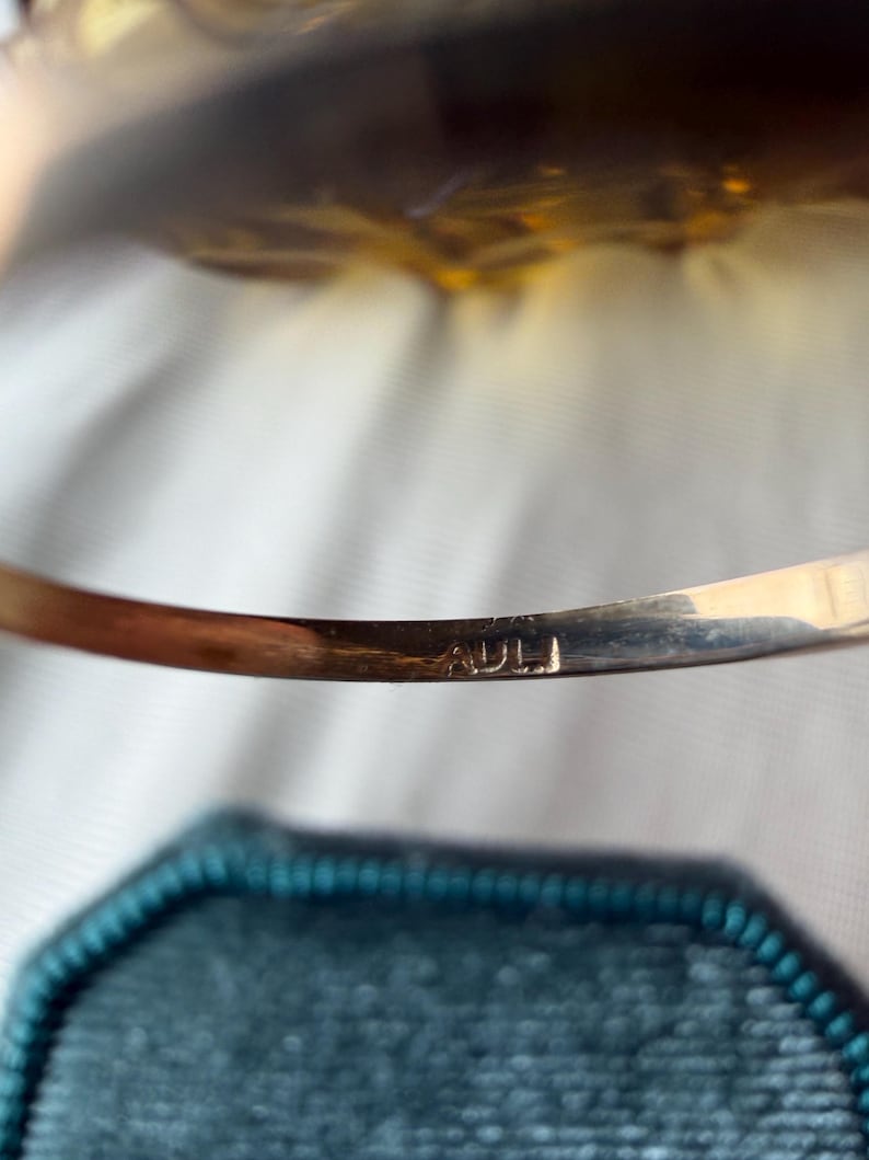 May include: A gold-toned bracelet with the engraved letters "ADLI" is displayed. The bracelet is slightly curved and rests above a teal, hexagonal jewelry box. The background is a soft, blurred white.